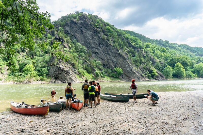A group of people are gathered on a rocky shore next to a river or lake, with several canoes pulled up onto the shore. Some individuals are standing near the canoes, while others are in the water. A large, rocky hill covered with trees rises in the background, and the sky is partly cloudy. The overall scene suggests an outdoor recreational activity, possibly canoeing or kayaking.